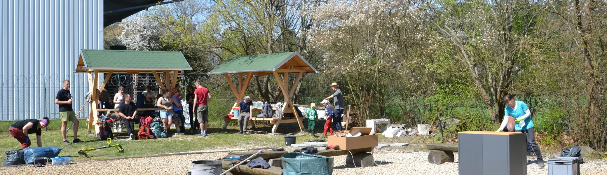 Personen ruhen sich unter den Holzhütten vom Arbeitseinsatz aus und unterhalten sich. | © Theodor Kubusch