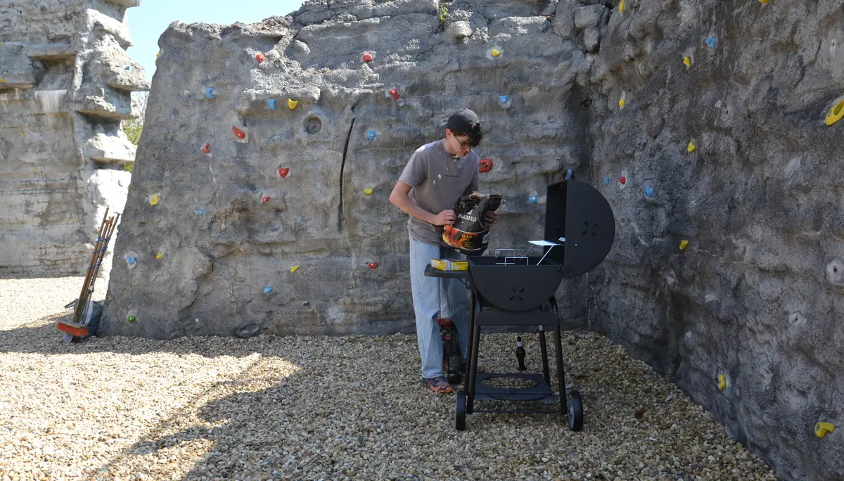 Eine Person kippt Holzkohle in den Grill; im Hintergrund die Boulderwand. | © 	 Theodor Kubusch