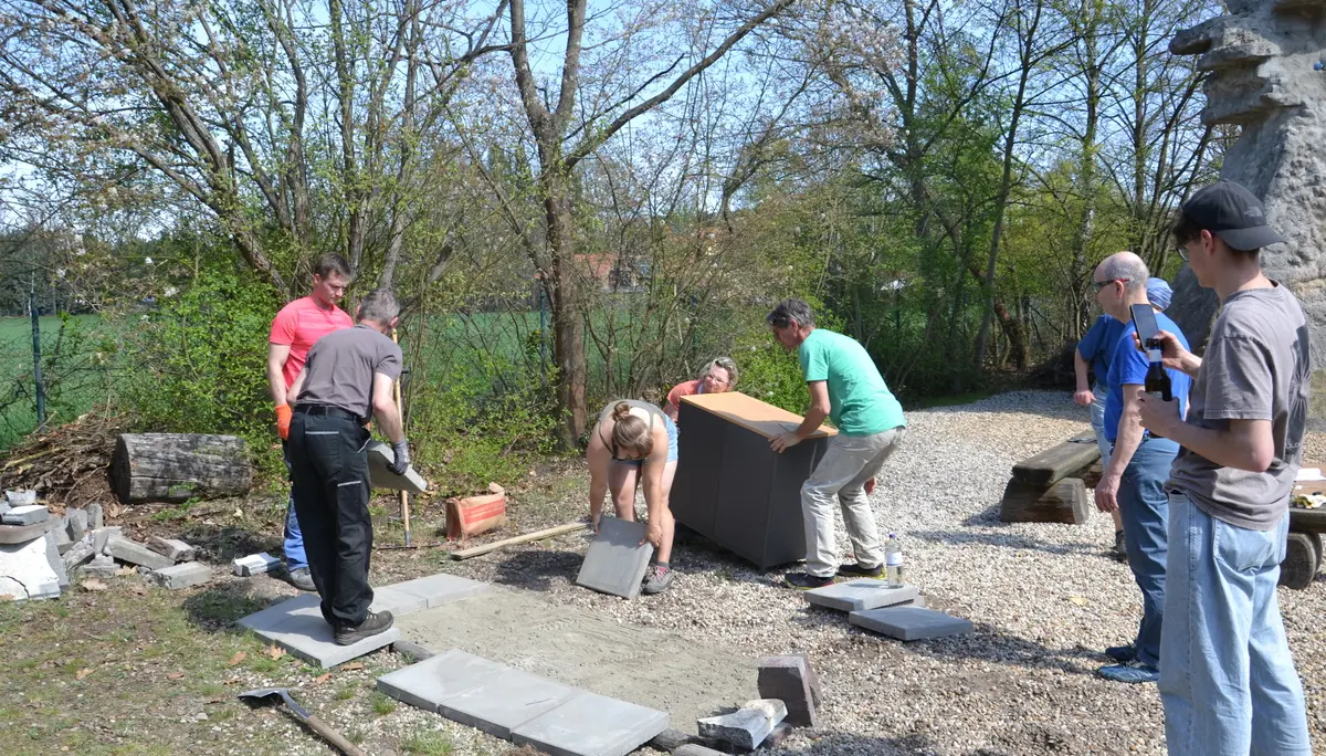 Mehrere Personen bereiten den Untergrund für den Grillplatz vor. Eine Person hebt eine Gehwegplatte, dahinter tragen zwei Personen den Grillschrank. | © 	 Theodor Kubusch