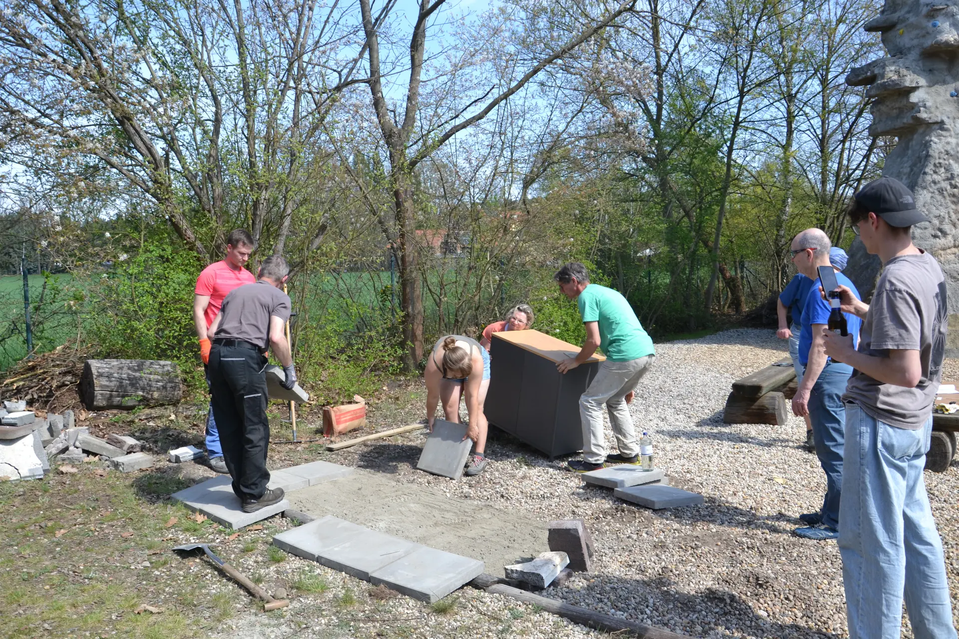 Mehrere Personen bereiten den Untergrund für den Grillplatz vor. Eine Person hebt eine Gehwegplatte, dahinter tragen zwei Personen den Grillschrank. | © 	 Theodor Kubusch